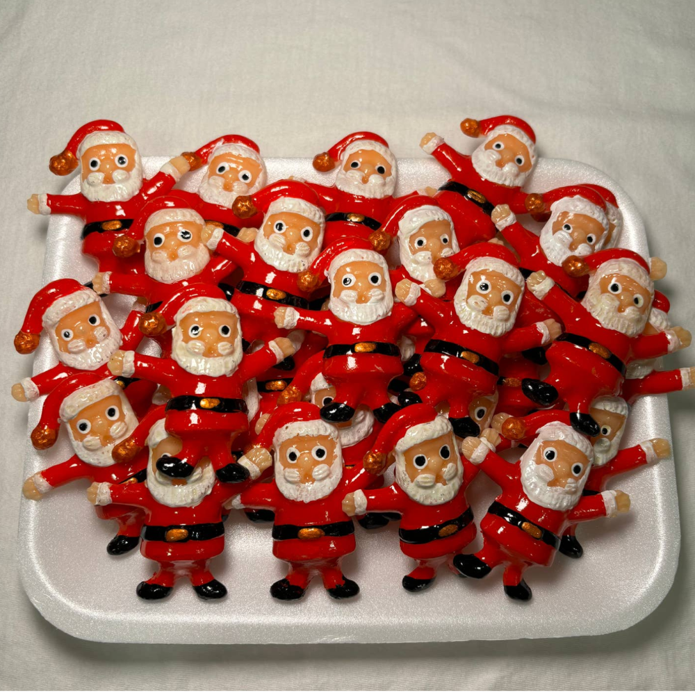 White tray full of clay Santa magnets. Each Santa magnet has a red body, black belt with gold buckle, white beard, and traditional red Santa hat