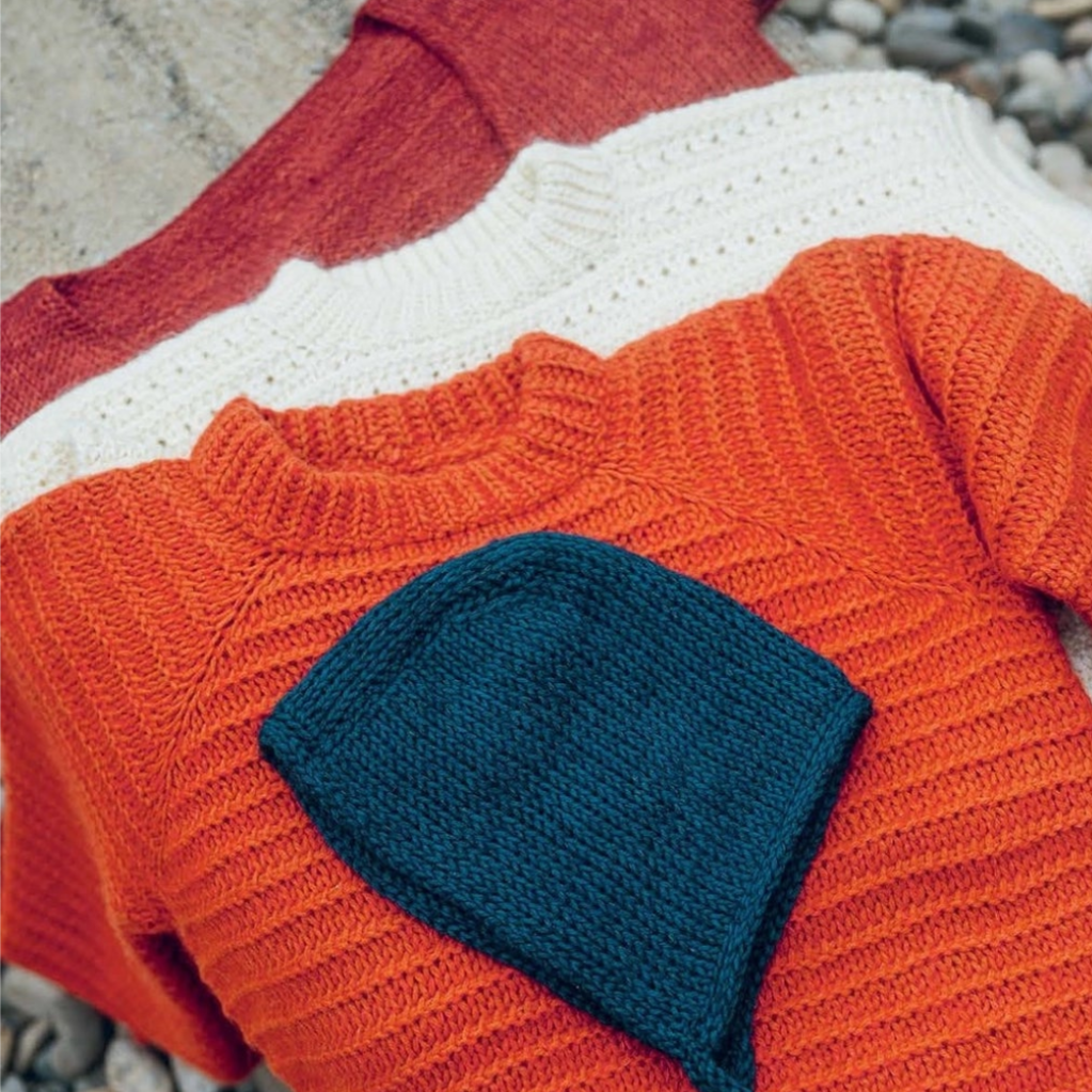 Knitted orange sweater with blue hat on pebbles, book content page on right