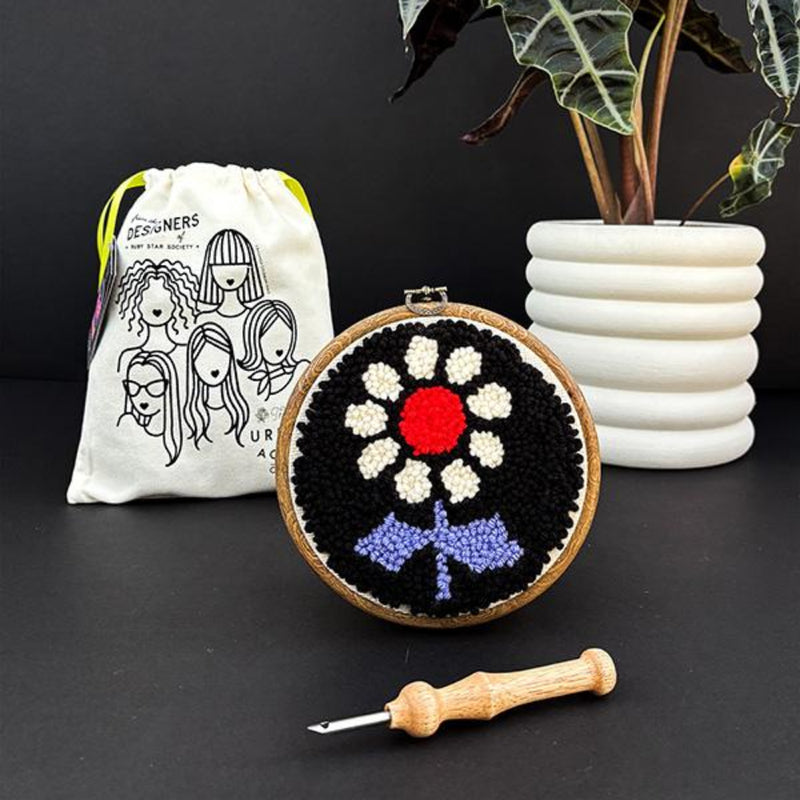 Embroidery hoop with floral design, white drawstring bag, plant, and tool on a dark surface