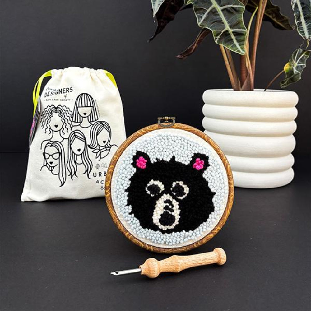 Embroidery hoop with a black and white bear design, surrounded by a drawstring bag and plant on a dark surface.