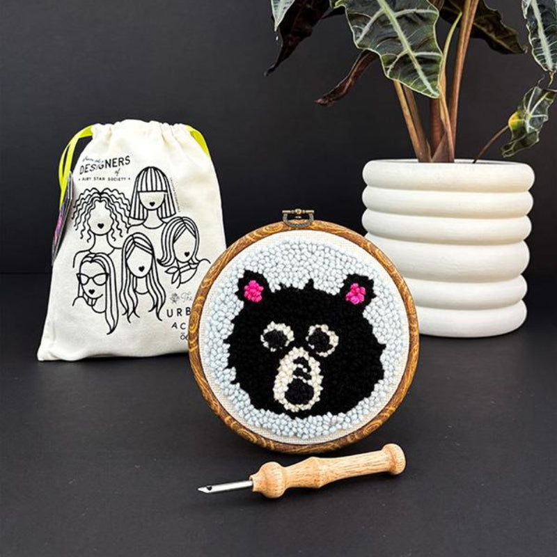 Embroidery hoop with a black and white bear design, surrounded by a drawstring bag and plant on a dark surface.