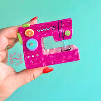 Pink sewing machine hair claw held by fingers with red nail polish on an aqua blue background.