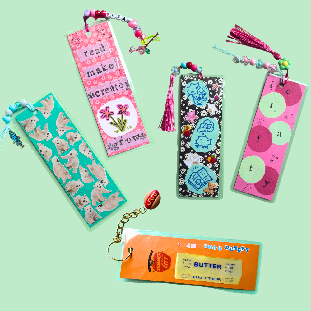 Five colorful bookmark-style items arranged on a pale mint background: a turquoise bookmark with dog illustrations, a pink bookmark with text and flowers, a black bookmark with blue cartoon motifs and tassels, a pink bookmark with circular letters and tassels, and an orange keychain-style tag with a butter ticket graphic.