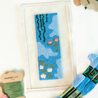 Needlepoint bookmark with a blue waterlily design on white fabric, surrounded by yarn and tools.