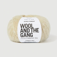 A ball of Wool and the Gang's Alpaca Stardust yarn in Stardust Gold, featuring a white paper label with black text, set against a clean, light gray background.