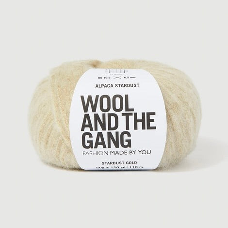 A ball of Wool and the Gang's Alpaca Stardust yarn in Stardust Gold, featuring a white paper label with black text, set against a clean, light gray background.