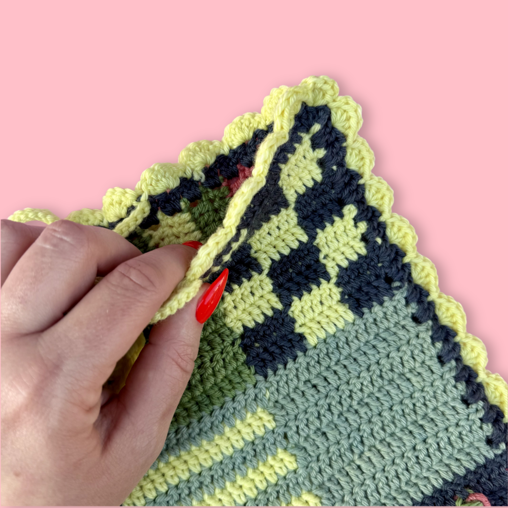 Hand holding a crocheted fabric with a checkered pattern on a pink background