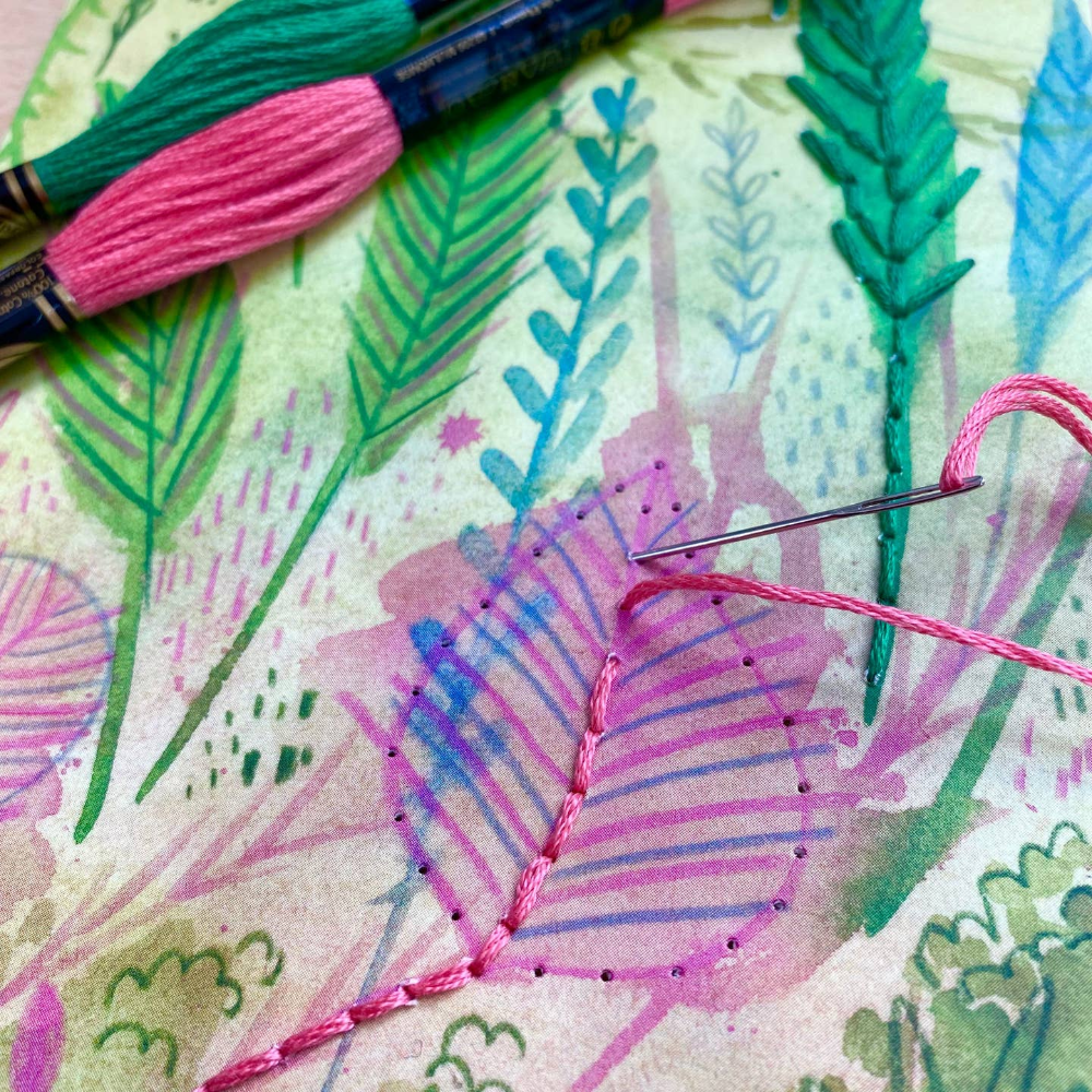 Fabric with floral pattern and pink thread, surrounded by embroidery tools.