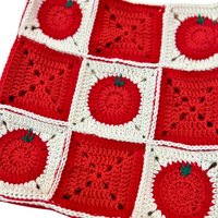 A close-up of a crocheted bag featuring alternating red and white square panels, with several tomato motifs and small green leaf details.