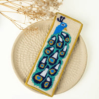 Needlepoint peacock bookmark on a textured surface with dried flowers.