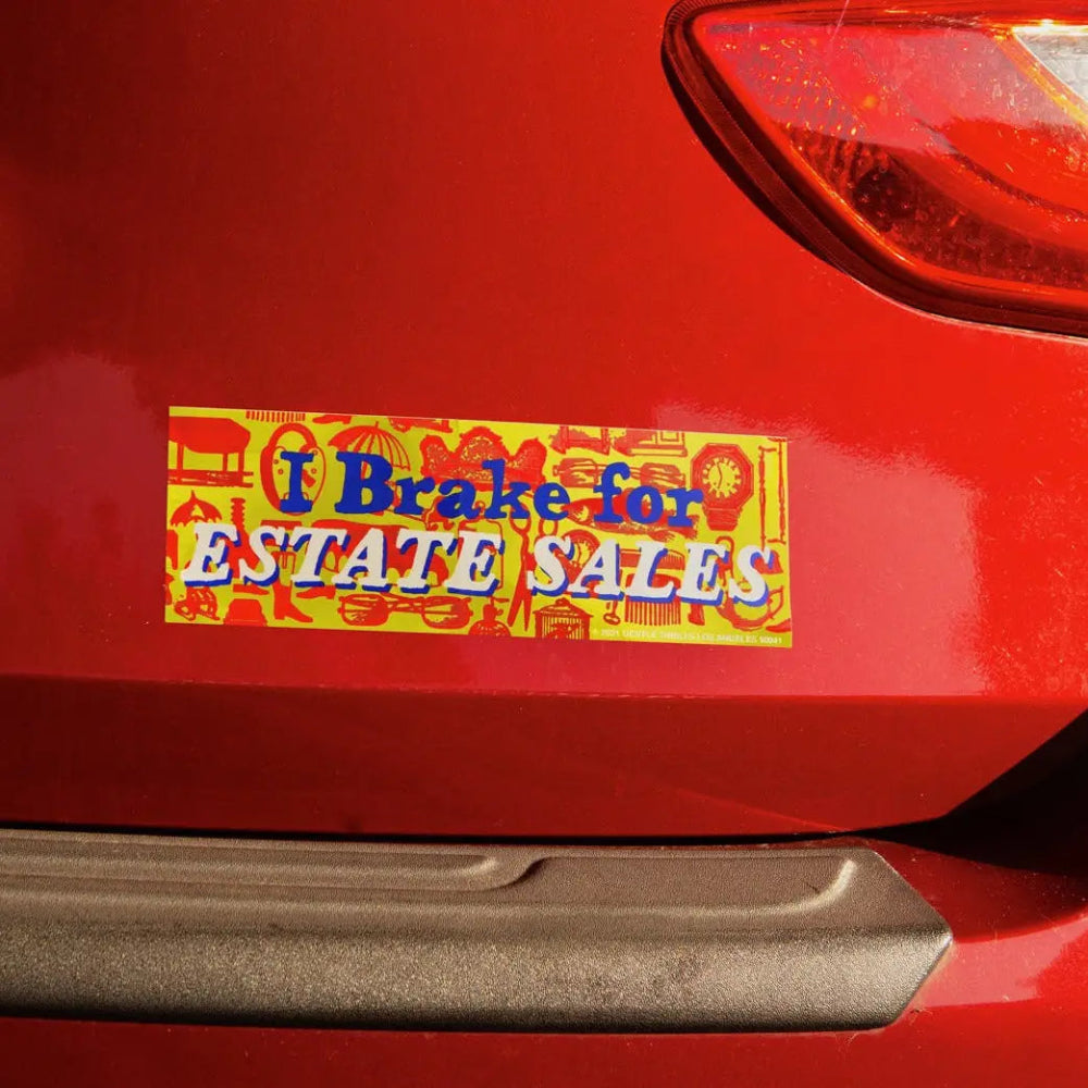 Bumper sticker on a red car reading 'I Brake for Estate Sales'.