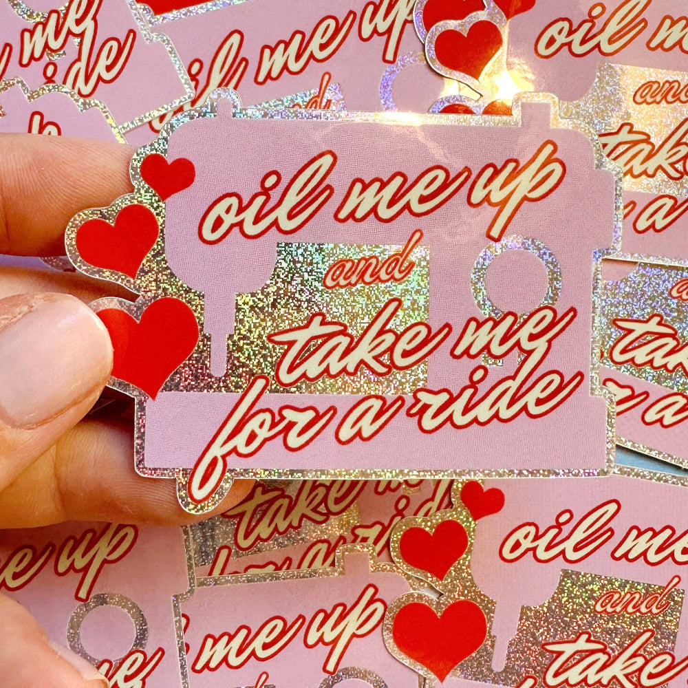 Sticker with text 'oil me up and take me for a ride' and red hearts, held by a hand.