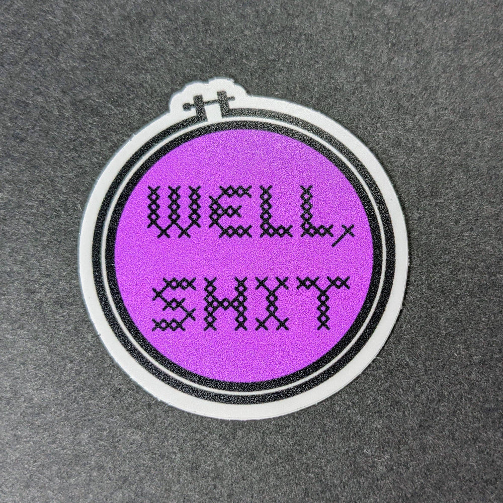 Embroidery hoop sticker with 'WELL SHIT' text on a purple background, placed on a textured gray surface.