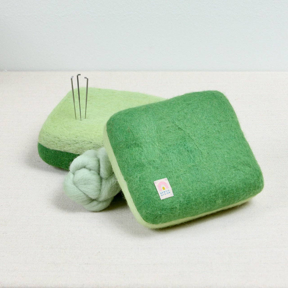 Green wool needle felting mat with green fiber and needle felting needles on a white background