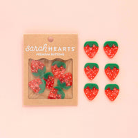 Strawberry-shaped buttons with a 'Sarah Hearts' packaging on a pink background