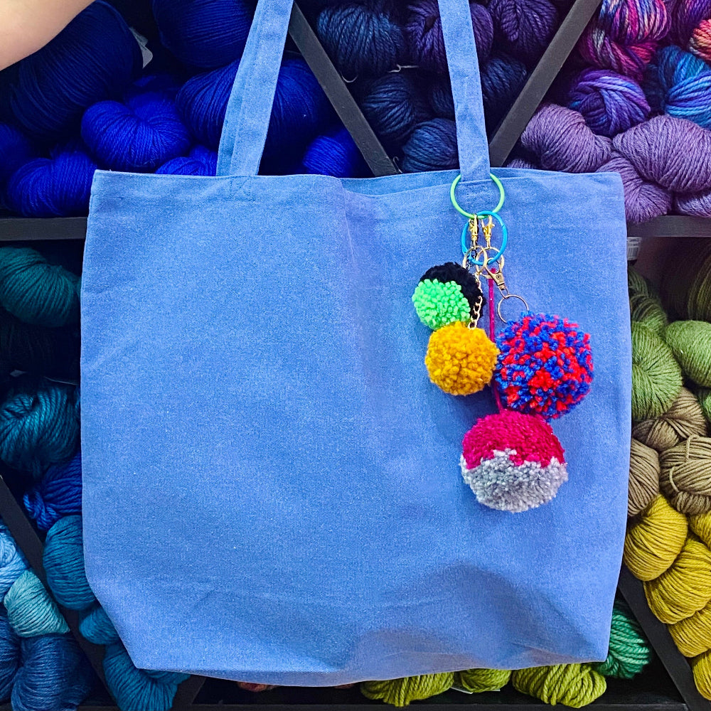 Blue tote bag with colorful pom-pom keychain against a backdrop of yarn.