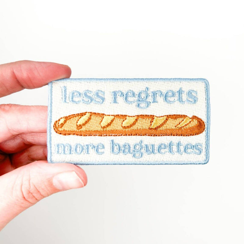 Patch with 'less regrets more baguettes' text held by a hand on a white background