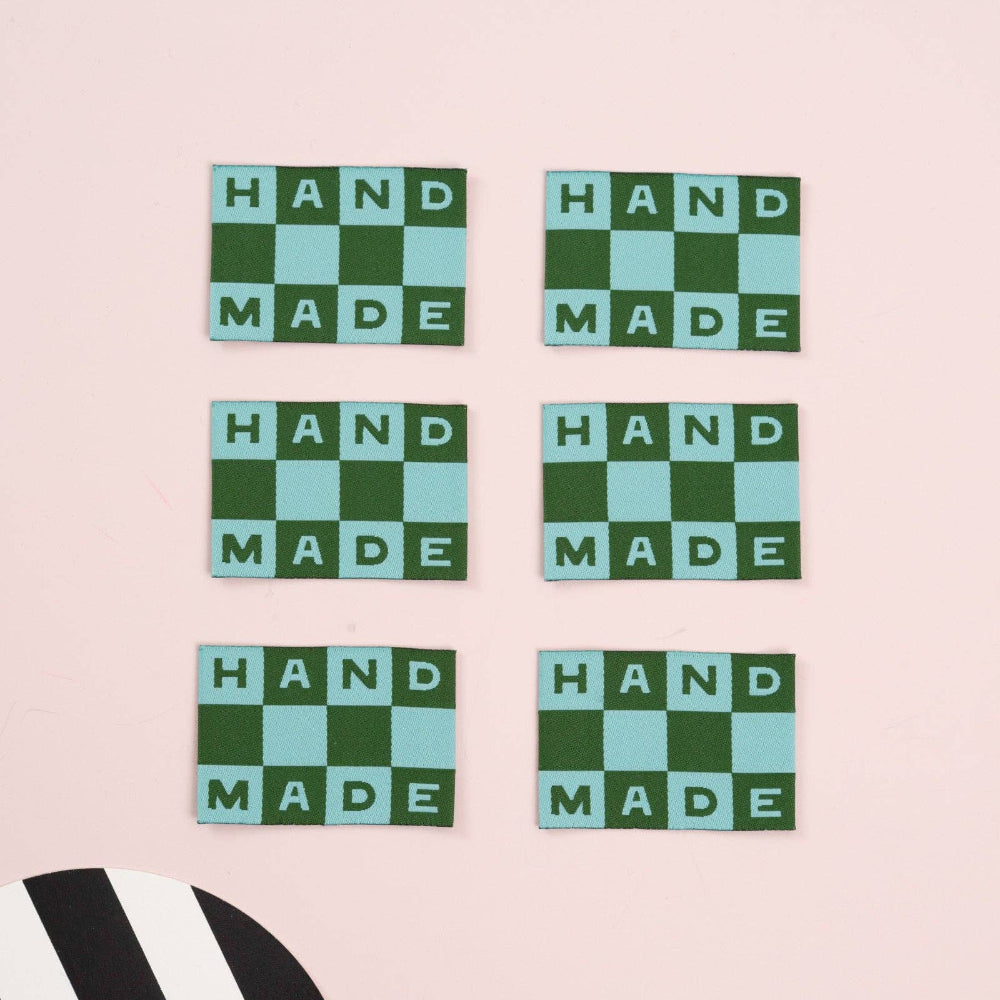 Five green and blue 'Hand Made' labels on a pink background with a black and white product at the bottom.