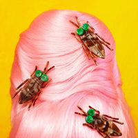Three decorative cockroaches with green eyes on a pink hairpiece against a yellow background
