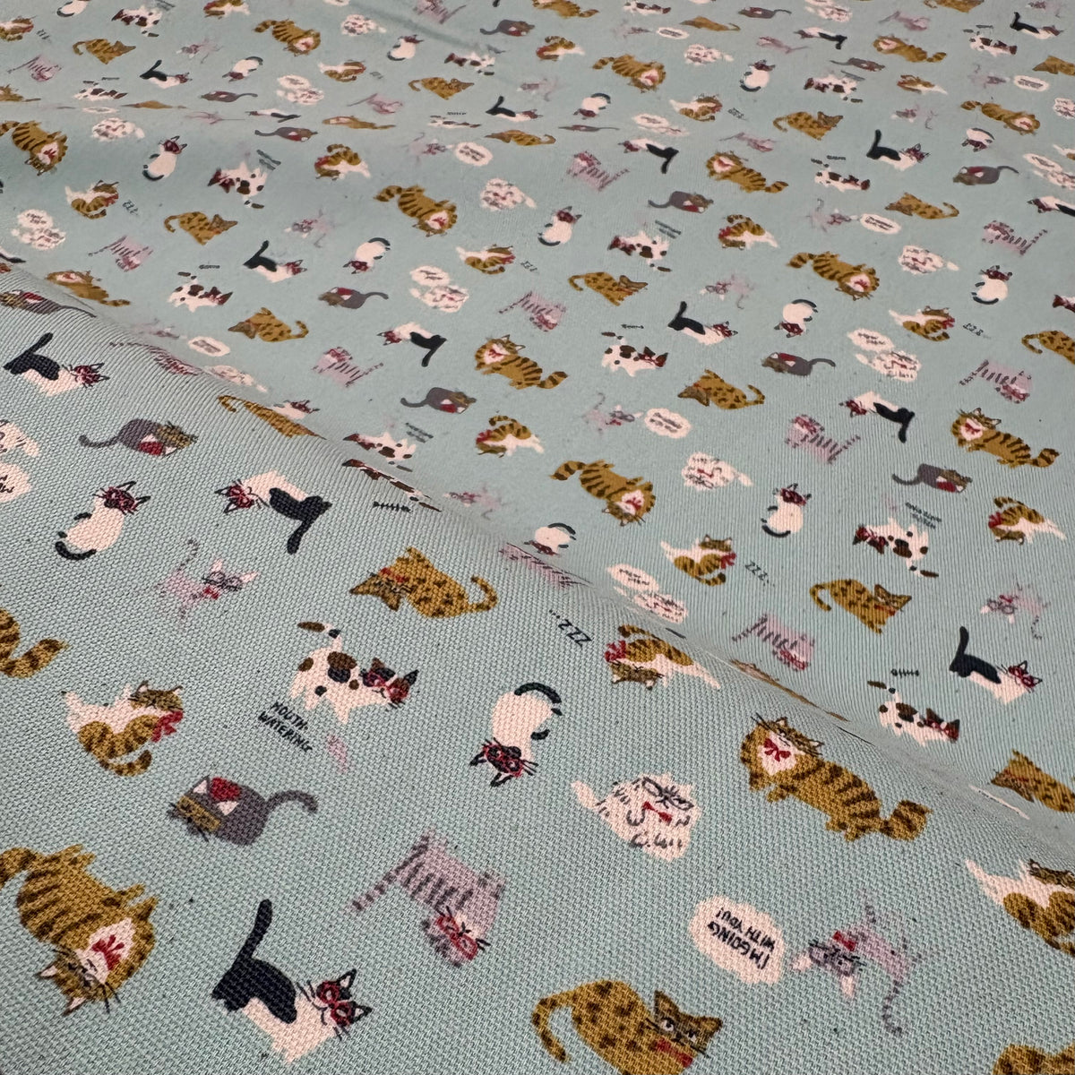 Close up of a bolt of light blue fabric featuring illustrations of cats wearing glasses and bowties.