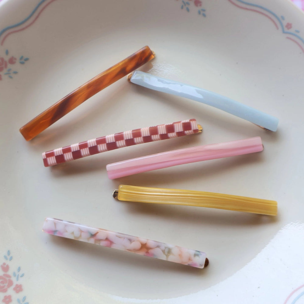 Six colorful hair claw clips arranged on a white ceramic plate with a pink floral border, set against a pink and white checkered tablecloth.