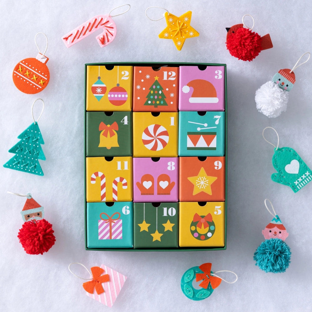 A colorful Christmas advent calendar with 12 numbered compartments surrounded by festive handmade ornaments on a white snowy background.