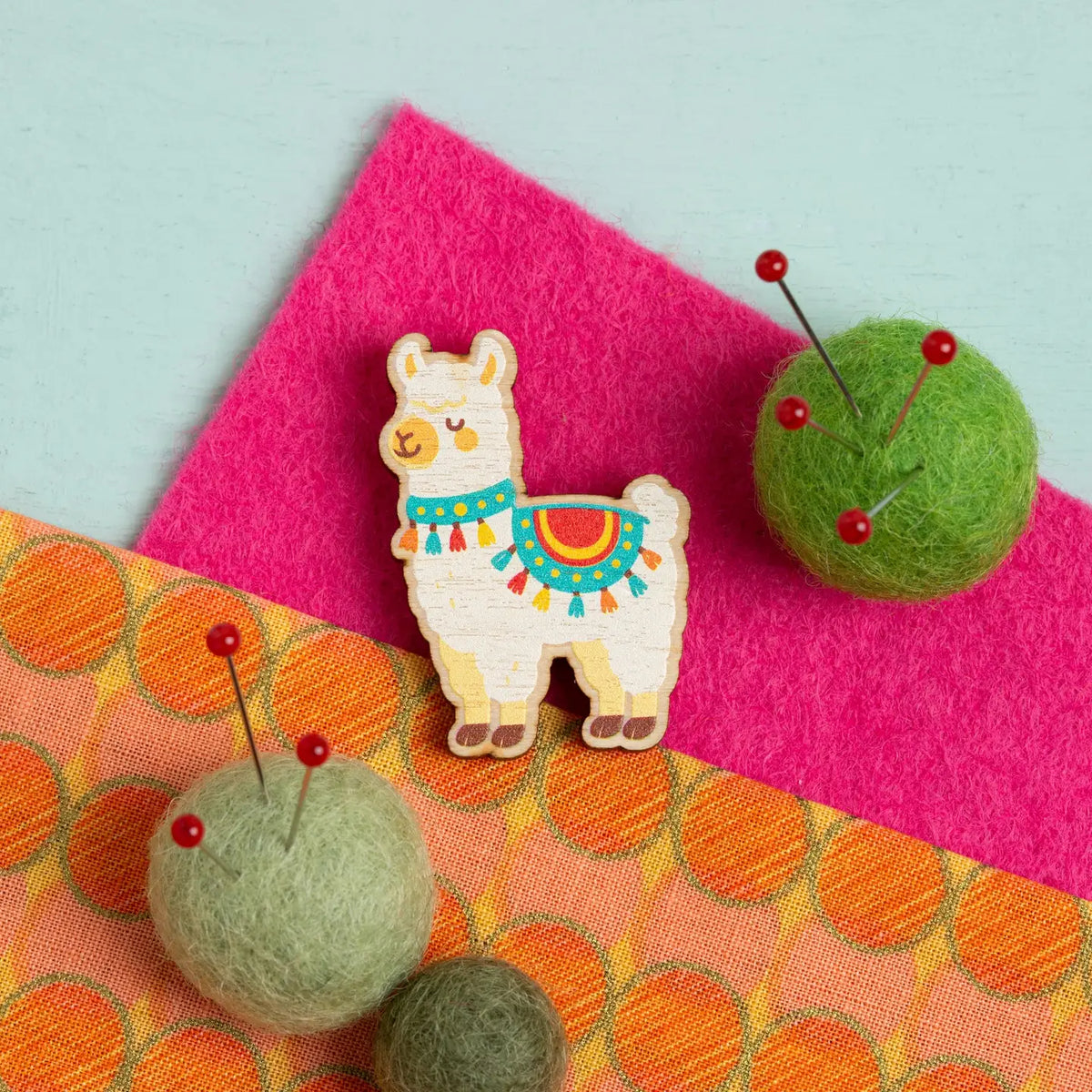 Colorful alpaca-shaped needle minder on a pink and orange fabric background with green felt balls with pins in them
