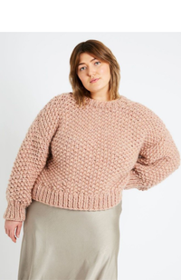 Brunette woman wearing a taupe satin skirt and an oversized blush pink knit sweater on a white background.