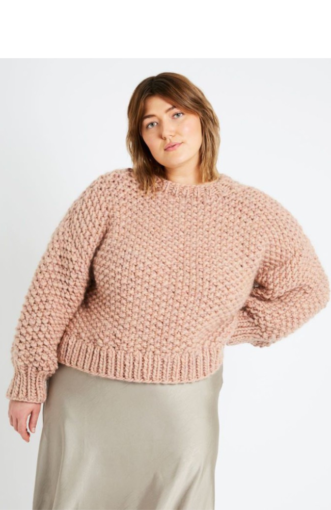 Brunette woman wearing a taupe satin skirt and an oversized blush pink knit sweater on a white background.