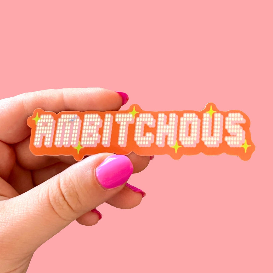 Hand with pink nail polish holdiing a sticker that reads "ambitchous," in a glossy glitter font resembling a disco ball and orange bored. Background is a solid light pink.
