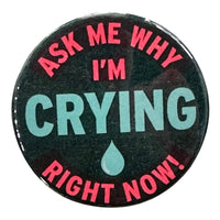A black button against a white background. Text on the button reads "Ask Me Why I'm Crying Right Now" in red text with the word crying and a tear drop in the center in blue. The surrounding text is in bright pink.