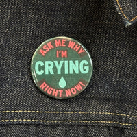 A button is placed on a denim jacket near the collar for size comparison. Text on the button reads "Ask Me Why I'm Crying Right Now" in red text with the word crying and a tear drop in the center in blue. The surrounding text is in bright pink. 