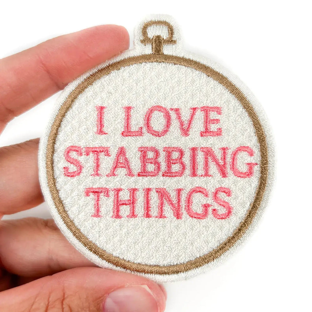 Embroidery hoop with 'I love stabbing things' text held by a hand on a white background