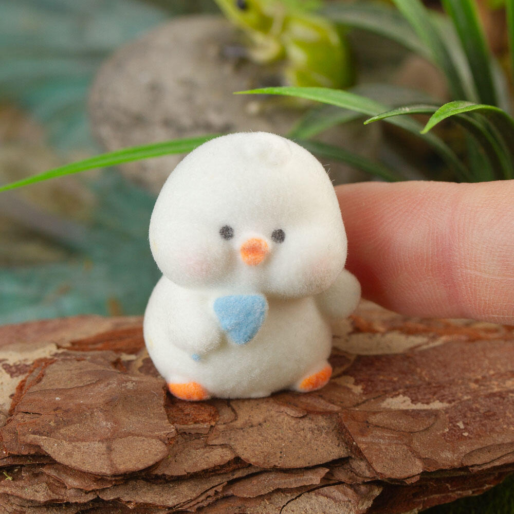 Small white flocked chick with a blue shovel near a person's finger, surrounded by green plants and wood.