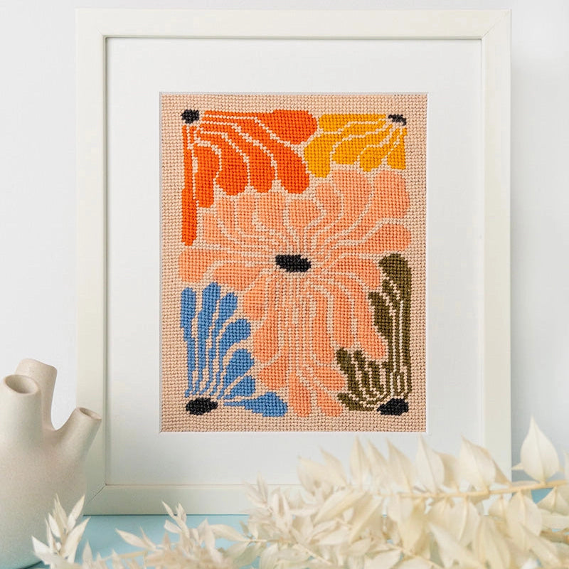 Framed needlepoint art with orange, blue, pink, and green flowers on a light blue surface with a white background