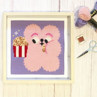 Cross-stitched pink bear with popcorn on a wooden surface with sewing supplies.