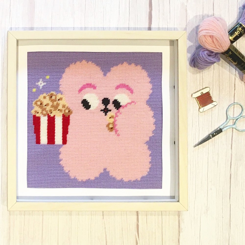 Cross-stitched pink bear with popcorn on a wooden surface with sewing supplies.