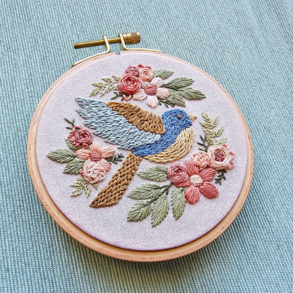 An embroidery hoop with a textured bluebird and elegant flowers embroidered on a pale pink background