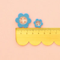 Two blue daisy-shaped buttons with pearl white centers and pink dots, shown above a yellow  scalloped edge ruler on a pale pink background.