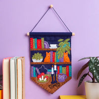 Colorful felt pennant featuring bookshelves, cats, plants, candles, mushrooms, glasses, and a coffee mug hanging on a purple wall.