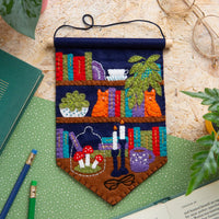 Colorful felt pennant featuring bookshelves, cats, plants, candles, mushrooms, glasses, and a coffee mug laying on a wooden table.