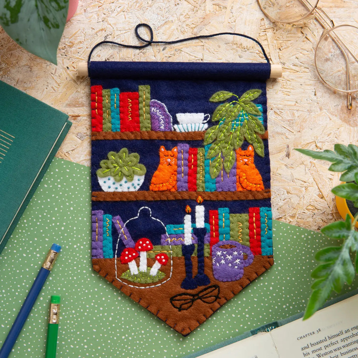 Colorful felt pennant featuring bookshelves, cats, plants, candles, mushrooms, glasses, and a coffee mug laying on a wooden table.