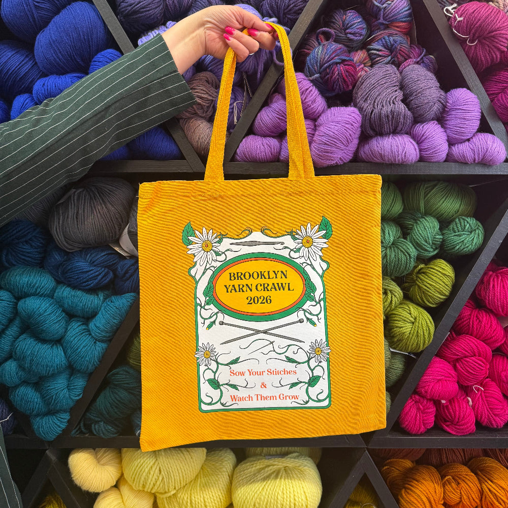 Yellow tote bag with 'Brooklyn Yarn Crawl 2026' design held in front of colorful yarn balls.