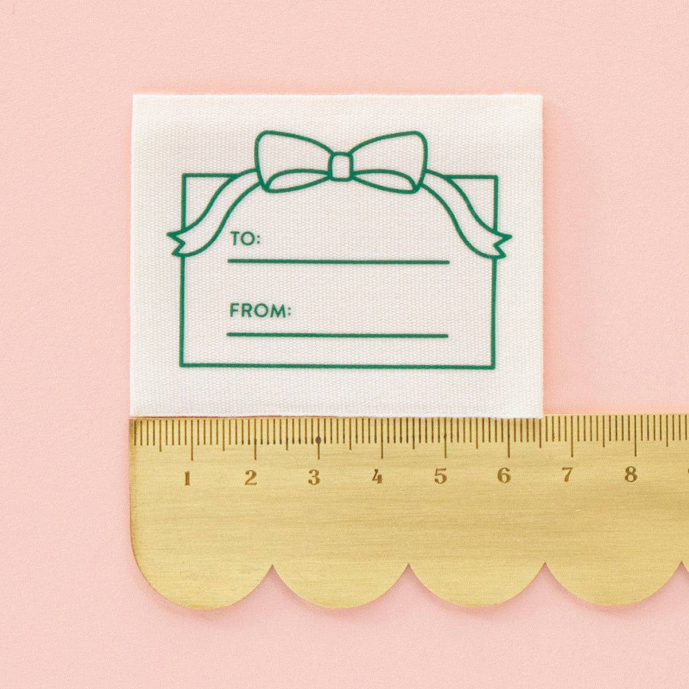 Gift tag label with a green bow design and 'TO' and 'FROM' fields, placed on a pink background with a gold ruler at the bottom.