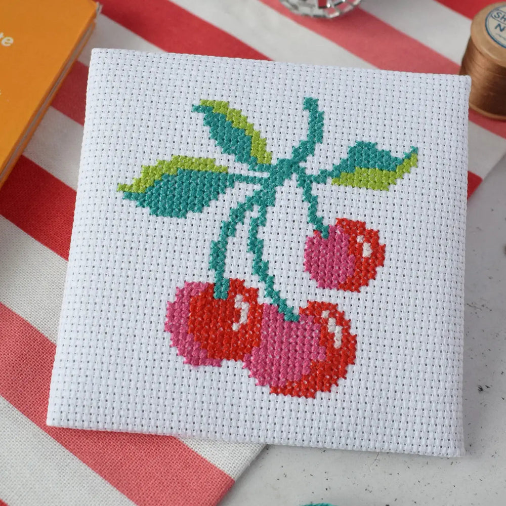 Cross-stitched cherry design on a white fabric square with a striped background