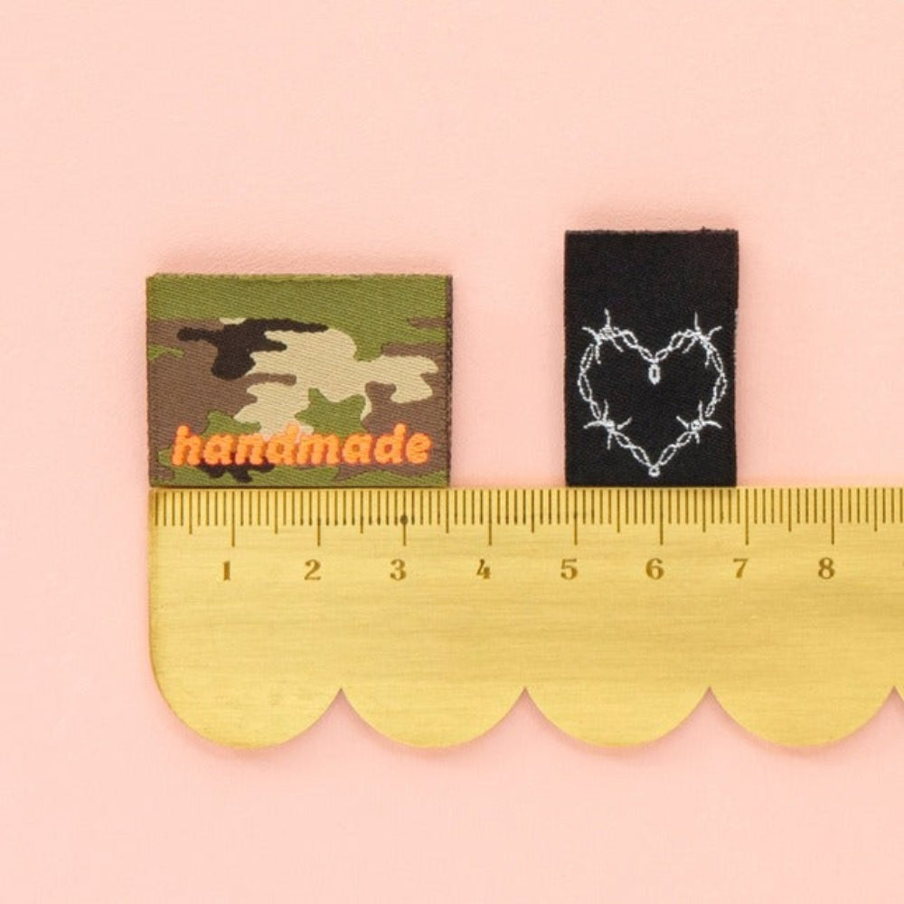 Two woven labels, one camouflage and one with a heart design, on a pink background with a ruler for scale.