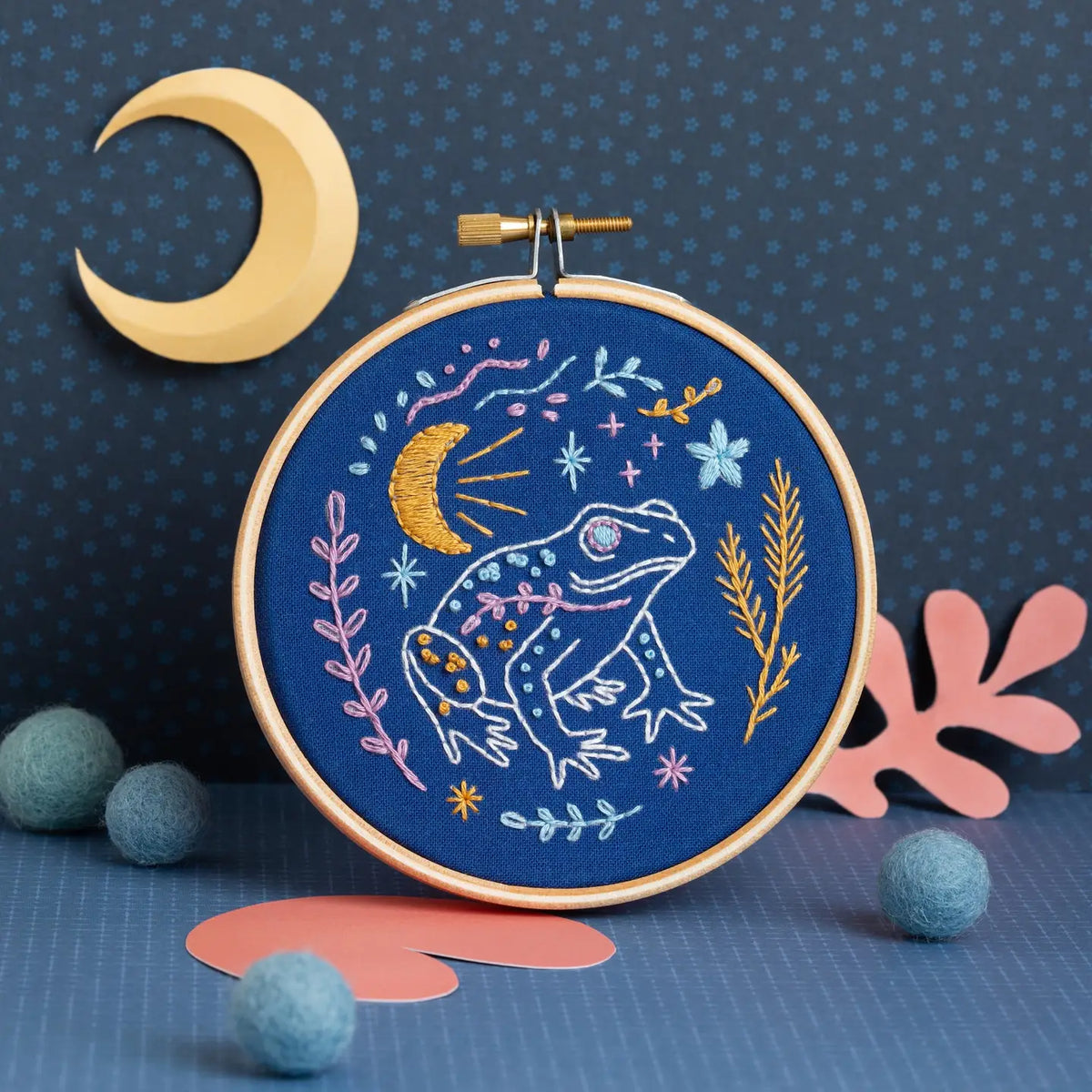 Embroidery hoop with a frog design on a blue background with decorative elements.