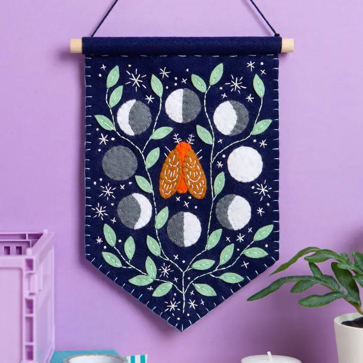Felt pennant featuring a moth, moon cycle, plants, and stars hanging on a purple wall