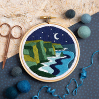 Embroidery hoop with a night scene of cliffs and moonlight embroidery, surrounded by felt balls and scissors on a textured surface.