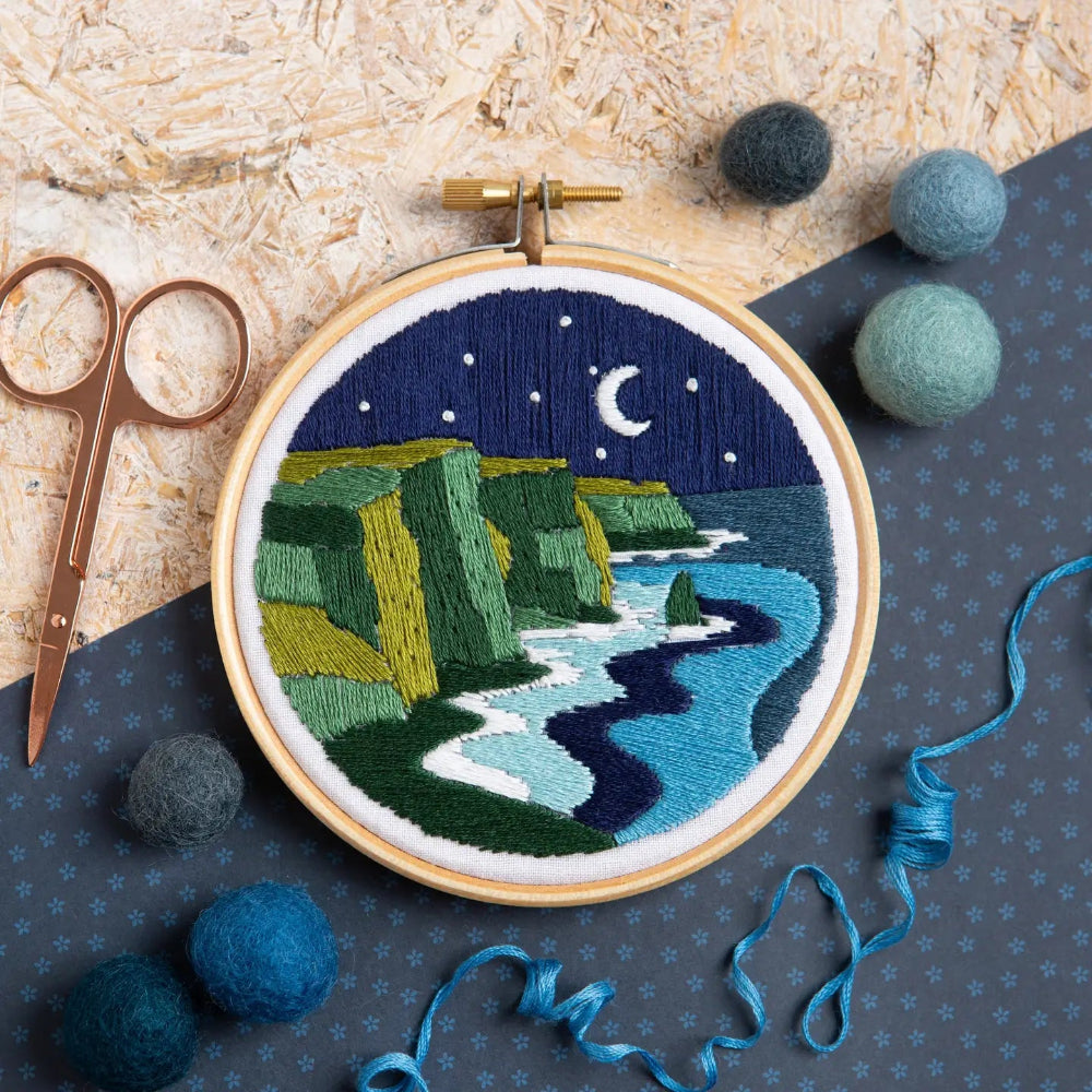 Embroidery hoop with a night scene of cliffs and moonlight embroidery, surrounded by felt balls and scissors on a textured surface.